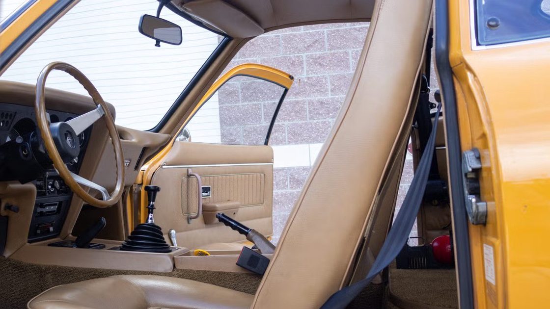 vehicle interior with steering wheel, manual gear shifter, beige seat, door panel controls.
