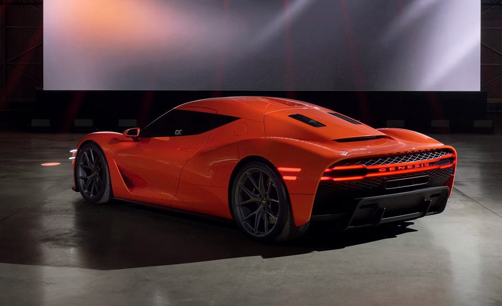 rear three quarter view of orange sports car on display stage with illuminated taillights and large digital screen.